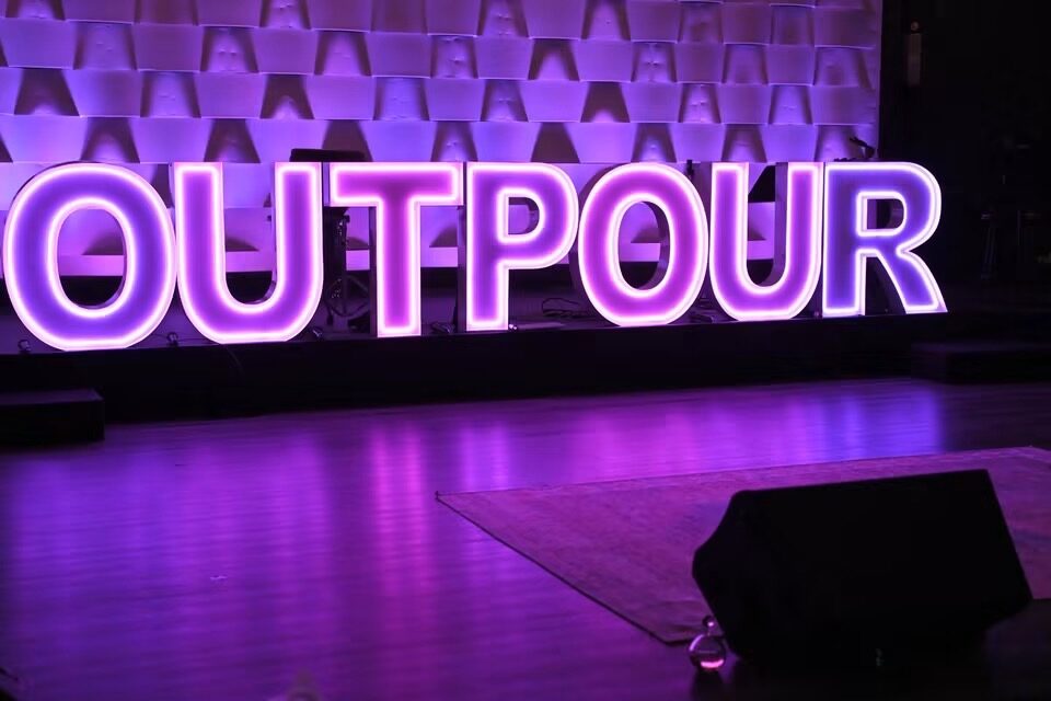 OUTPOUR in purple Illuminate letters on a lit stage