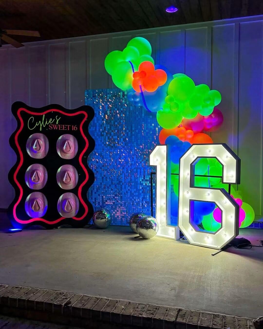 A lit 16 beside a neon cake sign and a green and orange balloon cluster