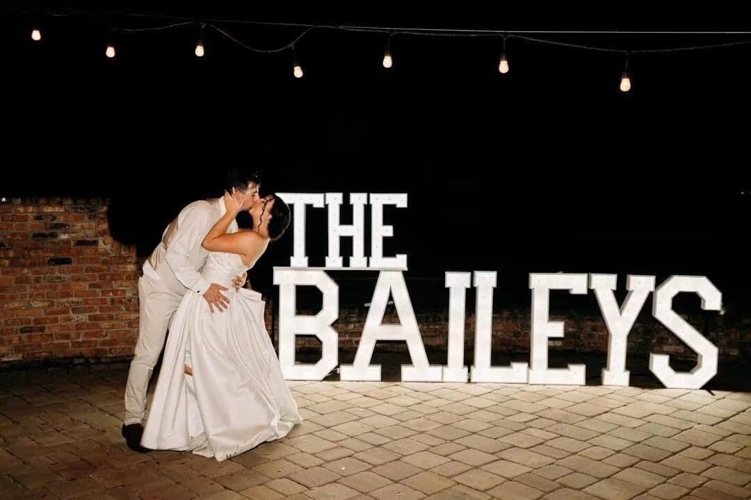 THE BAILEYS in lit letters under string lights with a bride and groom