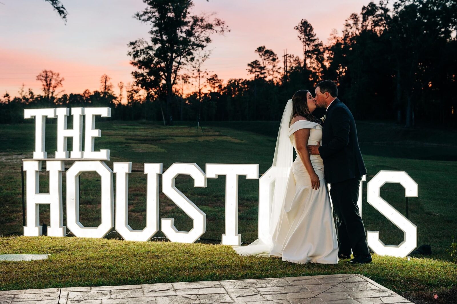 THE HOUSTONS in lit letters on a lawn at sunset behind a bride and groom