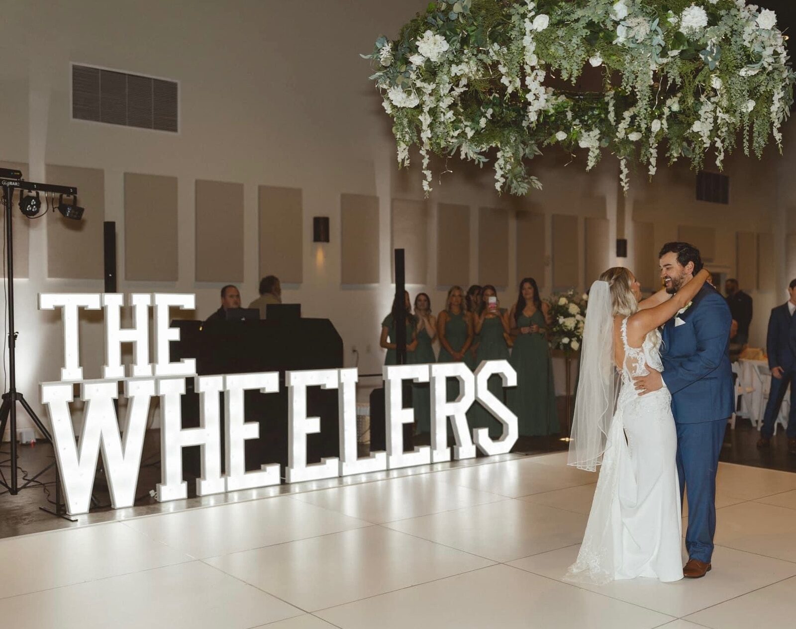 THE WHEELERS in lit letters below hanging florals at a reception
